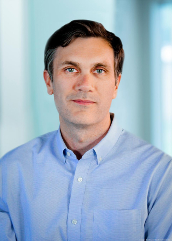 In the image, there is a man offering a subdued smile, appearing calm and approachable. He has short, dark hair, clear skin, and deep-set eyes. He's wearing a light blue, buttoned-up shirt that suggests a casual business style.