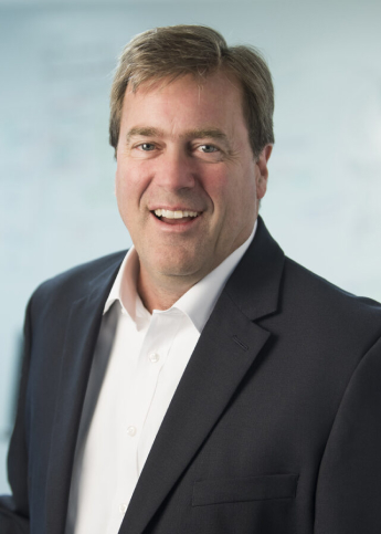 The photograph is a professional headshot of a man with a warm smile. He has medium-length brown hair and is wearing a dark business suit with a white shirt. The background is out of focus, suggesting a corporate or professional setting.