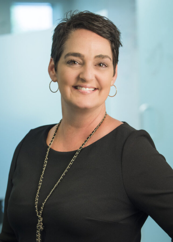 The image features a professional headshot of a woman with a confident smile. She has short, dark hair styled neatly, hoop earrings, and wears a black outfit complemented by a long necklace. The background is blurred, likely an office setting